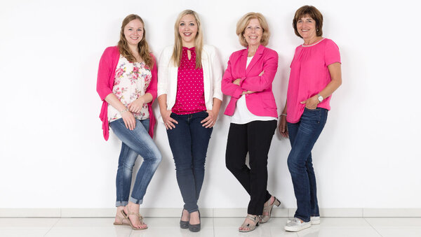 Vier Frauen in modischer Kleidung in Pink und Weiß stehen lächelnd vor weißem Hintergrund.